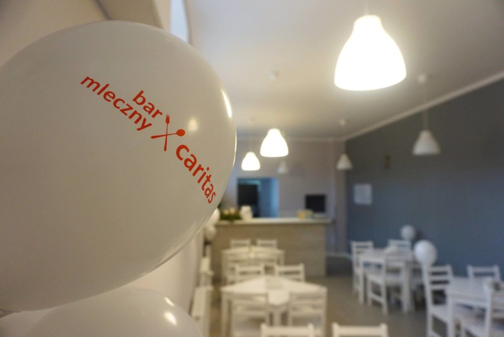 Zdjęcia ukazuje wnętrze lokalu gastronomicznego (stoliki z krzesłami, lada barowa), wnętrze jest utrzymane w jasnych kolorach. Na pierwszym planie widoczny jest biały balon, a na nim napis: bar mleczny caritas.
