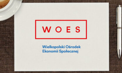 Widok z góry, całą powierzchnię zajmuje brązowe biurko a na nim na środku biała kartka, na niej logo WOES Wielkopolski Ośrodek Ekonomii Społecznej. Obok kartki leży długopis oraz filiżanka kawy.