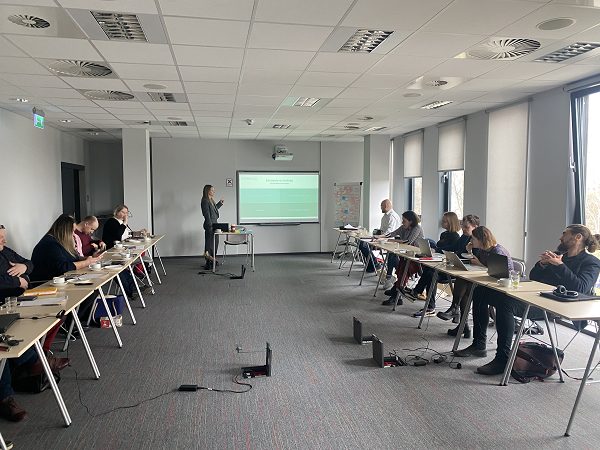 Na zdjęciu wnętrze sali konferencyjnej. Dwa rzędu stolików, przy której siedzą uczestnicy szkolenia. Na końcu sali Marta Szymborska wskazuje slajd na tablicy multimedialnej.